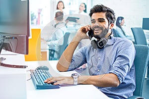 Smiling handsome man employee in business wear talking on mobile phone at his workspace at computer in office.