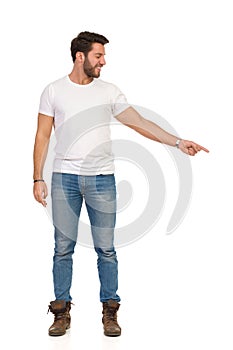 Smiling Handsome Man In Jeans And White T-shirt Is Standing And Pointing Down. Front View