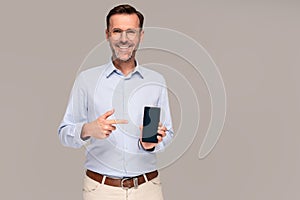 Smiling handsome man pointing on empty screen of smartphone, isolated on gray background