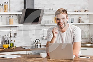 smiling handsome man working with laptop in kitchen and looking