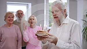 Smiling handsome senior man blowing out candles on birthday cake as friends singing congratulating clapping at