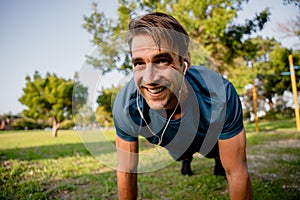 Smiling handsome young male athlete planking exercise in sunny outdoor park ear phones playing music