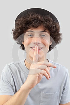 Smiling and handsome, the young man with blue eyes wears a hat and gestures with his finger to ask for silence, isolated against a