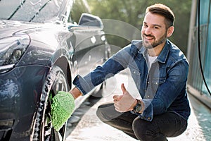 Smiling handsome young man showing thumb up and looking at camera, while using green wash mitt for cleaning rims of his