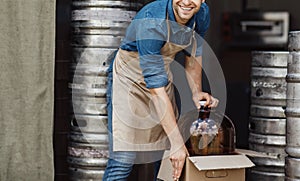 Smiling handsome young worker man in apron takes out large jar for brewing and production drinks