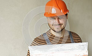 Smiling handyman repairman worker in overalls and protective orange helmet holds big putty knife for plastering concrete