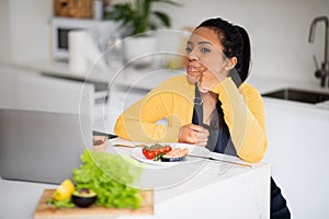 Smiling hangry young african american female in apron licks finger, looks at laptop and fresh fish, enjoys dish