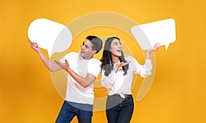 Smiling happy Asian couple holding blank speech bubbles on yellow background.