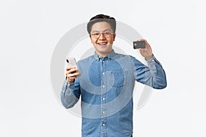 Smiling happy asian guy in glasses and casual clothes using mobile phone to transer money to bank account, showing