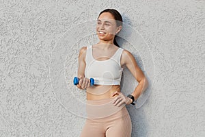 Smiling happy attractive dark haired woman of young age wearing white top and beige leggins, holding dumbbell in hands, doing