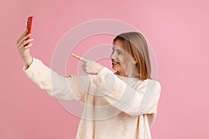 Smiling happy blond woman pointing to cell phone camera while having video call or livestream.