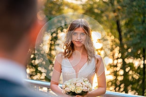 Smiling Happy Bride Holding Wedding Bouquet In Front Of Groom at Wedding Ceremony Outdoors At Park. Groom Is On