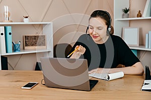 Smiling happy businesswoman in headphones sits at desk, looks at laptop screen, making notes, participating