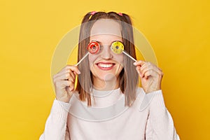 Smiling happy cheerful funky teenage girl with ponytails in jumper posing isolated over yellow background covering eyes with