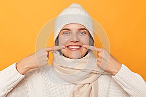 Smiling happy cheerful young caucasian woman wearing white sweater, hat and scarf, pointing at her teeth, showing result after