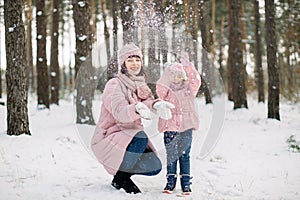 Smiling happy cute child daughter throwing snow up and having fun on a walk with her pretty loving mother. Mom and child