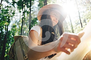 Smiling and happy cute traveler woman wearing backpack and hat hold location map in hands among trees at sunset