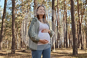 Smiling happy delighted Caucasian pregnant woman waking in spring forest wearing casual clothing rouching her big belly looking