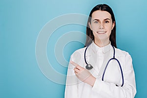 Smiling happy doctor pointing with finger on copy space, wear white lab coat stethoscope, isolated on blue studio background