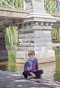 Boy sitting by lake in Boston public garden by bridge
