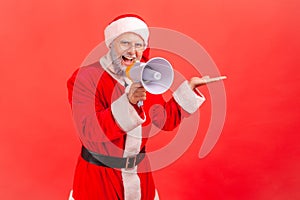 Smiling happy elderly man in santa claus costume showing freespace for advertisement and talking into loudspeaker, holidays