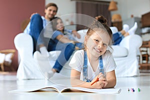 Smiling happy family sit relax on couch in living room watch little daughter drawing in album with colorful pencils