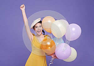 smiling happy fun young asian woman in yellow dress celebrating holding bunch of balloons raise up hand isolated on purple