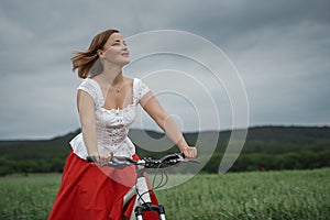 A smiling happy girl in a white blouse and red skirt rides a Bicycle on the edge of a wheat green field and does not look at the