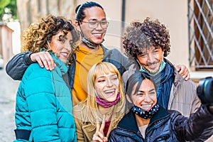 Smiling happy group of friends taking a selfie portrait with a reflex camera wearing warm winter clothes and protective face mask