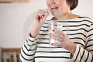 Smiling happy healthy senior woman at home holding glass of natural water taking daily pill for good health and senile