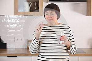 Smiling happy healthy senior woman at home holding glass of natural water taking daily pill for good health and senile