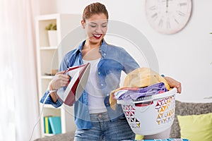 Smiling and happy housewife posing with iron and basket with clean laundry
