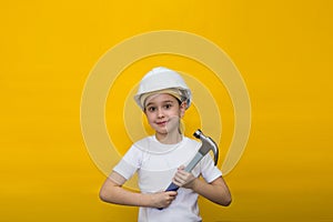 Smiling happy little girl in a construction white helmet holding a blue hammer on a yellow background