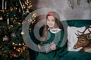 Smiling happy little girl in green dress by Christmas tree with glowing garland lights. Beautiful small girl in living room opens