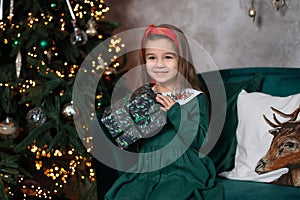 Smiling happy little girl in green dress by Christmas tree with glowing garland lights. Beautiful small girl in living room opens
