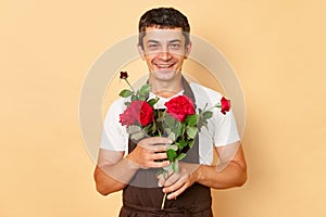 Smiling happy male florist wearing brown apron standing isolated over beige background at flowers shop embracing beautiful red