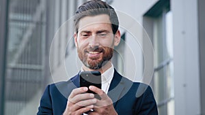 Smiling happy man holds phone browsing social network standing outdoors successful businessman responding to business or