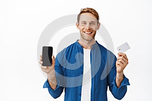 Smiling happy man showing mobile phone blank screen and bank credit card, recommending smartphone application, online