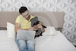 Smiling happy man sitting on the bed and hugging his lovely pet black pug breed and working on laptop
