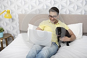 Smiling happy man sitting on the bed and hugging his lovely pet black pug breed and working on laptop