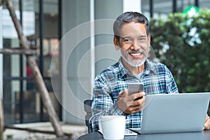 Smiling happy mature asian man with white stylish short beard using smartphone gadget serving internet at coffee shop cafe outdoor