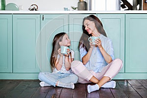 Smiling happy mother and daughter sitting against green cupboard, dreaming together