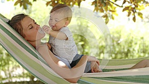 Smiling happy mother hugs and cuddle her little baby son, relaxing  lying together in the hammock. Child plays with pacifier, in