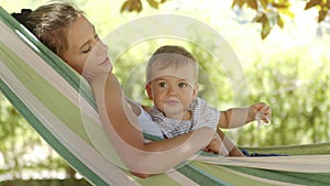 Smiling happy mother hugs and cuddle her little baby son, relaxing  lying together in the hammock. Child plays with pacifier, look