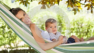 Smiling and happy mother hugs her little baby son, lying down relaxing in the hammock, in the home garden on a beautiful and sunny