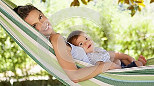 Smiling and happy mother hugs her little baby son, lying down relaxing in the hammock, in the home garden on a beautiful and sunny