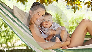 Smiling and happy mother hugs her little baby son, lying down relaxing in the hammock, in the home garden on a beautiful and sunny