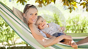 Smiling and happy mother hugs her little baby son, lying down relaxing in the hammock, in the home garden on a beautiful and sunny