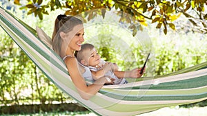 Smiling and happy mother hugs her little baby son, using smartphone, lying down relaxing in the hammock, in the home garden on a