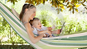 Smiling and happy mother hugs her little baby son, using smartphone, lying down relaxing in the hammock, in the home garden on a